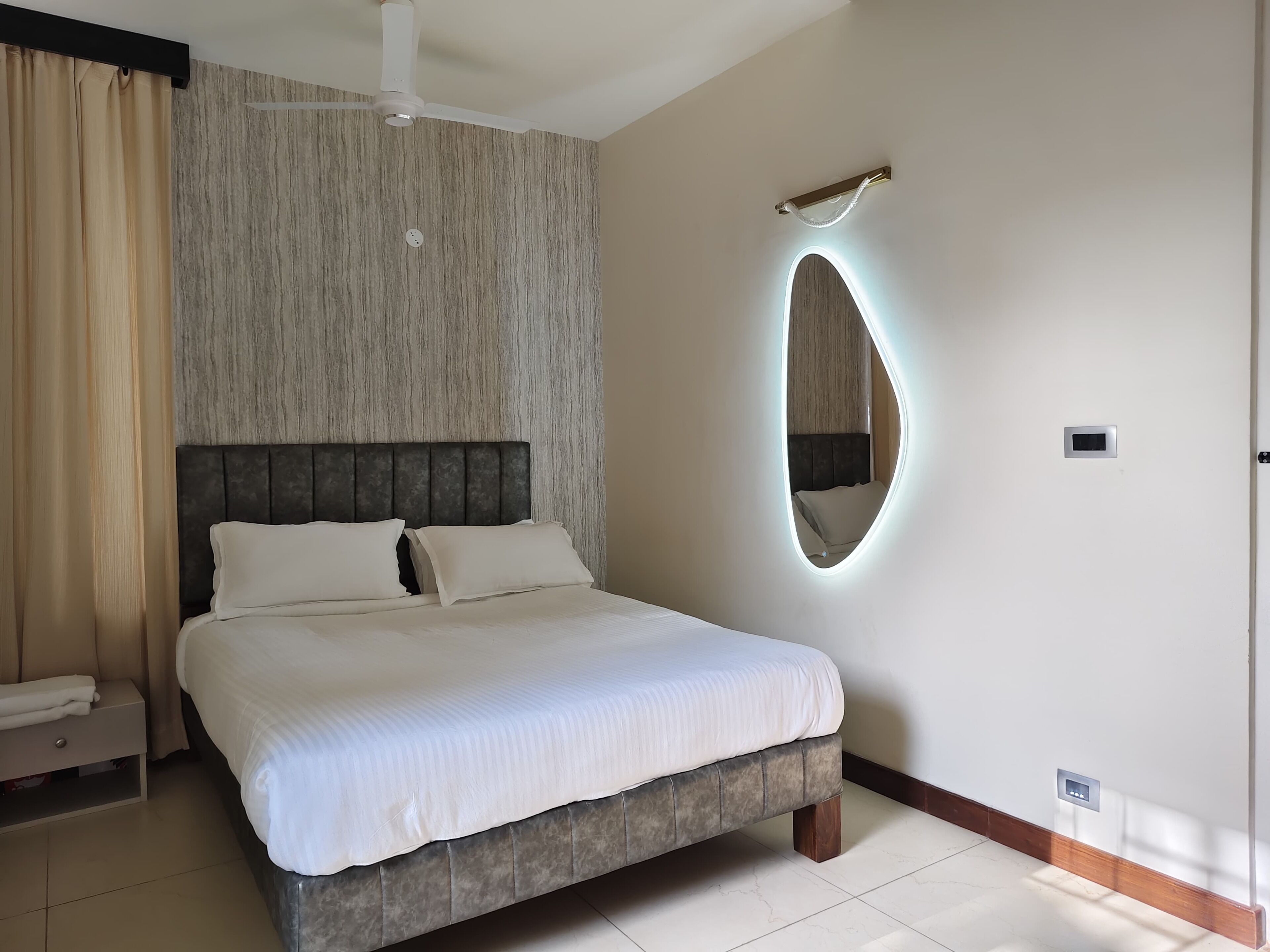 A neatly made bed with a headboard, a bedside table with a towel, a wall-mounted mirror, and a ceiling fan.
