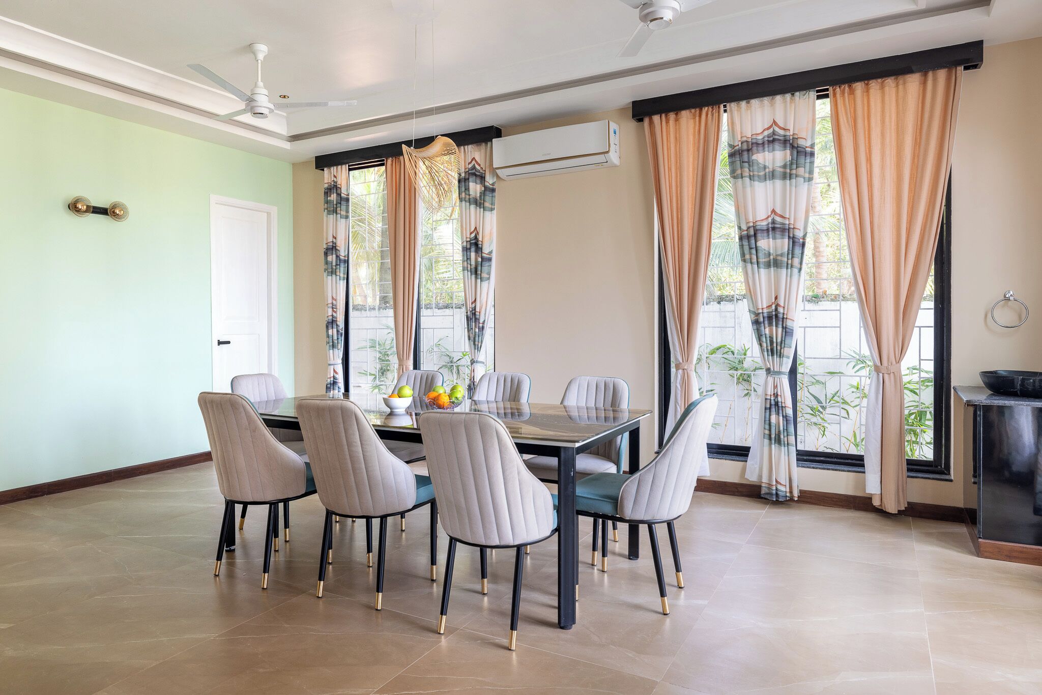 Un espace repas comprenant une table et des chaises, un ventilateur de plafond et de grandes fenêtres avec des rideaux.