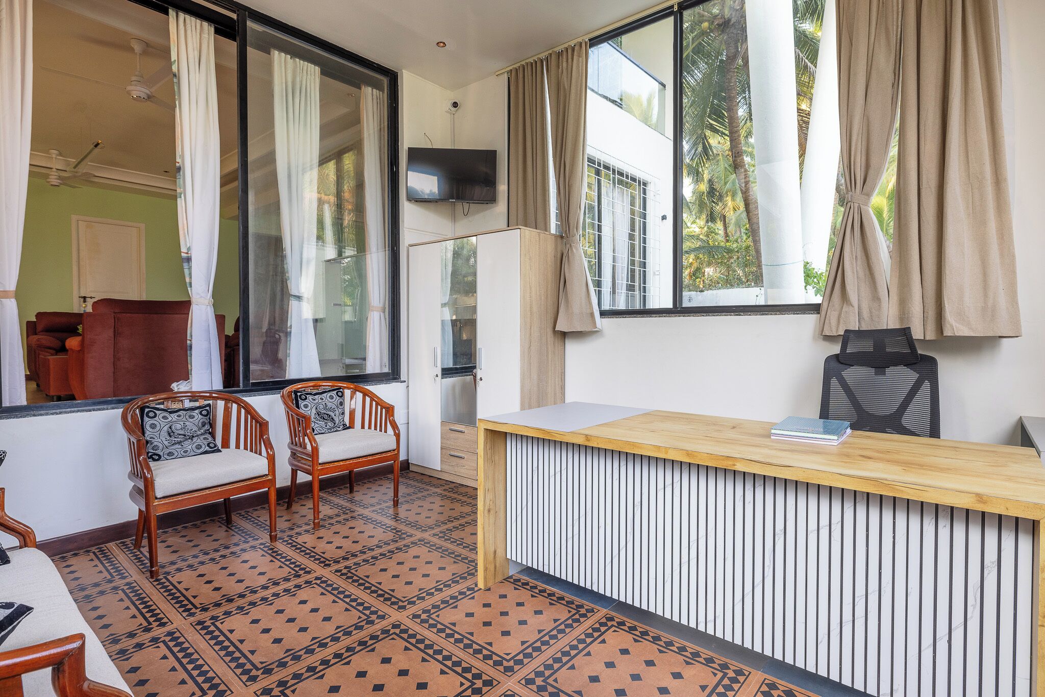Una habitación moderna con un escritorio de madera, dos sillas, un televisor y ventanas amplias con cortinas.