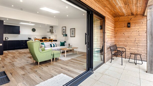 A modern interior with a wooden ceiling, a kitchen with dark cabinets, a green sofa, and a dining area with a glass table. - Home Farm Holiday Homes (Nuneham Courtenay)