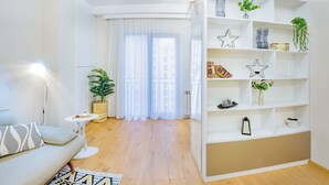 A modern living room with a sofa, a small table, a bookshelf, and a large window with sheer curtains.