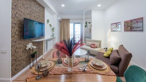 A modern living room with a dining area, a sofa, a TV mounted on the wall, and a decorative vase with plants.