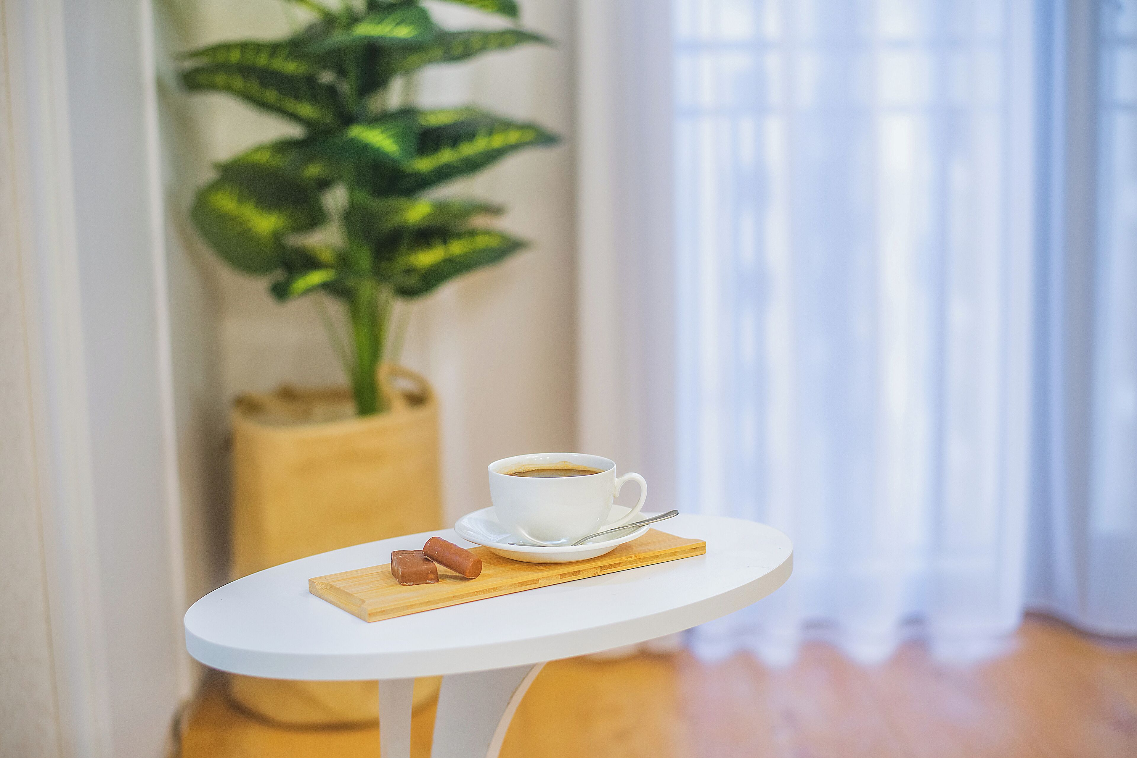 Una taza de café blanco sobre una pequeña mesa blanca con una bandeja de madera, una planta y una ventana con cortinas al fondo.
