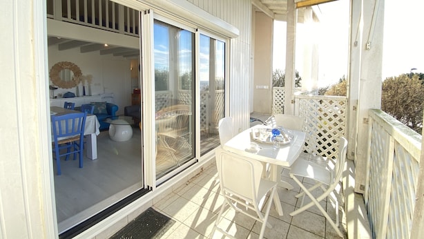 Uma área externa coberta com mesa e cadeiras, porta de vidro deslizante que dá para um espaço interno com mesa de jantar e cadeiras, e um cercado branco.