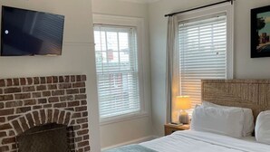 Ein Schlafzimmer mit Kamin aus Ziegelsteinen, einem Bett mit blauer Decke, einem Deckenventilator und einem an der Wand befestigten Fernseher.