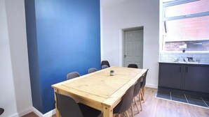 A room with a wooden dining table, black chairs, a blue accent wall, a white door, and a kitchen area with dark cabinets and a window.