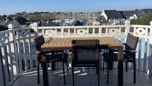 En terrasse med et træbord og stole, hvid gelænder og udsigt til huse og både i baggrunden.