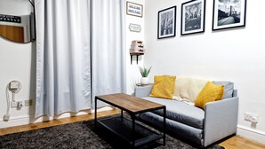 Una sala de estar moderna con un sofá gris, una mesa de centro de madera y una alfombra negra. Hay tres cuadros enmarcados en la pared, una planta en maceta y un espejo.