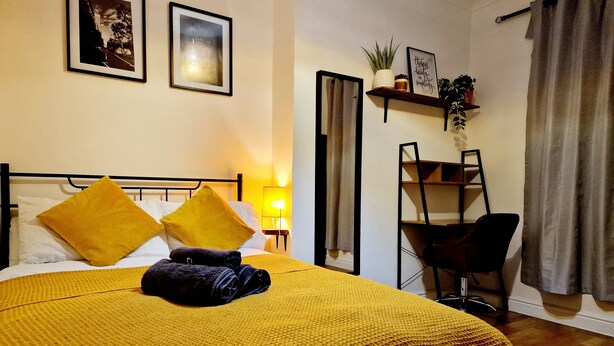 Una cama bien hecha con ropa de cama y almohadas amarillas, una lámpara de noche, un estante con plantas y una silla en una habitación con piso de madera y una ventana con cortinas.