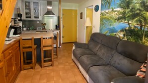 Una sala acogedora con un sofá gris y una zona de comedor con sillas de madera y una mesa. Se ve una cocina con armarios blancos y una estufa al fondo.