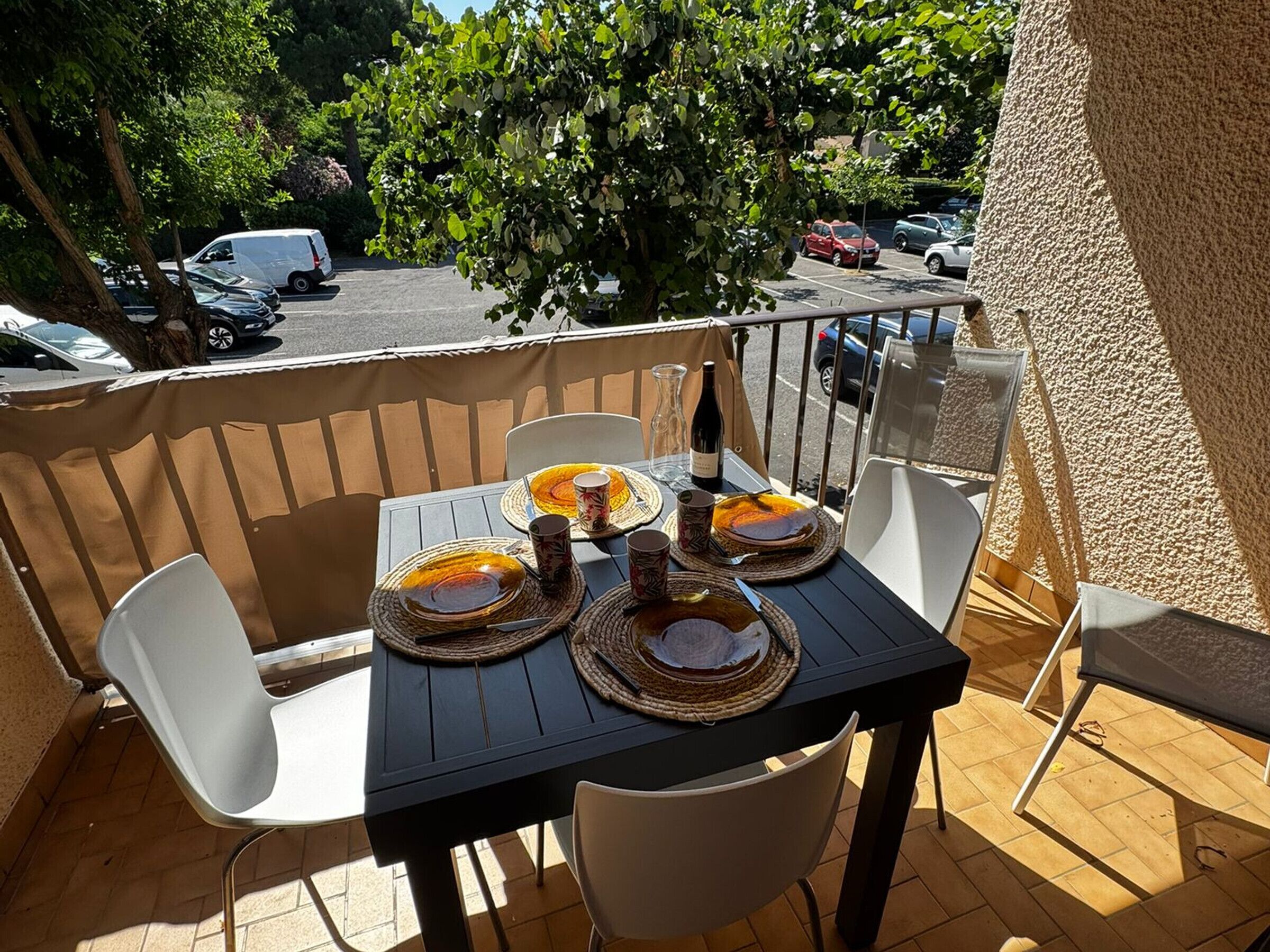 An outdoor dining area with a table set for two, surrounded by chairs, on a balcony with a view of parked cars and trees.
