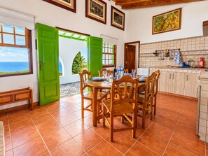 Un coin repas avec une table dressée pour quatre personnes, une cuisine avec un mur recouvert de carreaux, et une vue sur la mer par une fenêtre.
