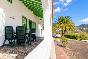 Un bâtiment à la façade blanche et aux touches de vert, une terrasse avec des sièges extérieurs, et une vue sur un paysage vallonné parsemé de palmiers.