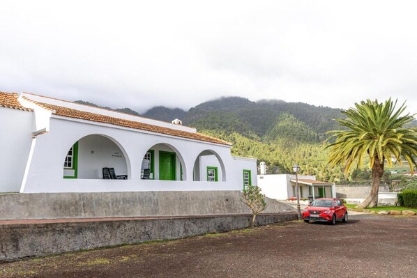 Un edificio blanco con puertas y ventanas verdes, un coche rojo estacionado al frente y una palmera.