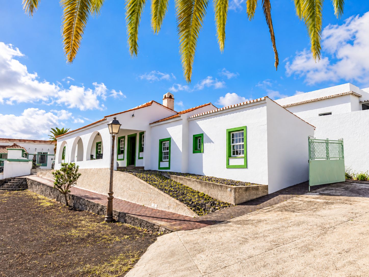 A white building with green window frames and a tiled roof, surrounded by a paved area and a small garden.