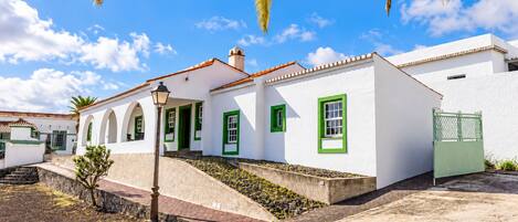 A white building with green window frames and a tiled roof, surrounded by a paved area and a small garden.