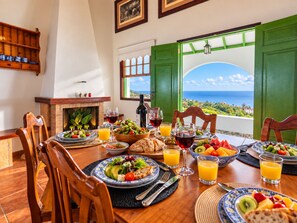 Mesa de comedor preparada con platos de comida, vasos de vino y jugo, y vista al océano a través de puertas verdes abiertas.