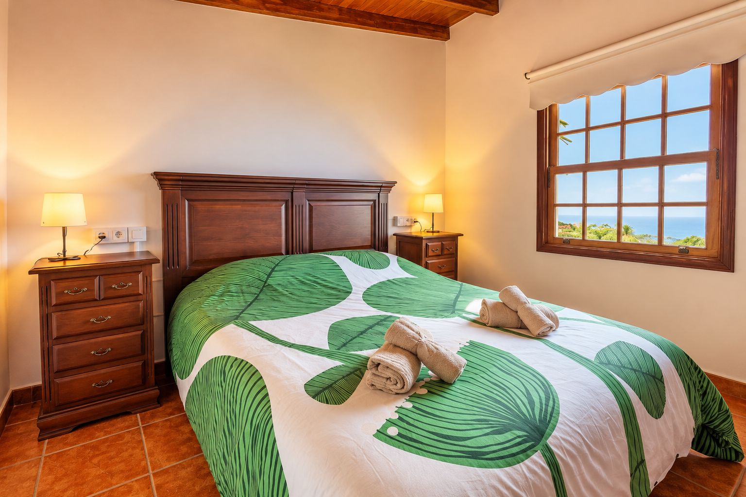 Habitación de hotel con una cama grande, cabecera de madera, dos mesitas de noche con lámparas, una ventana con vistas y suelo de baldosas.