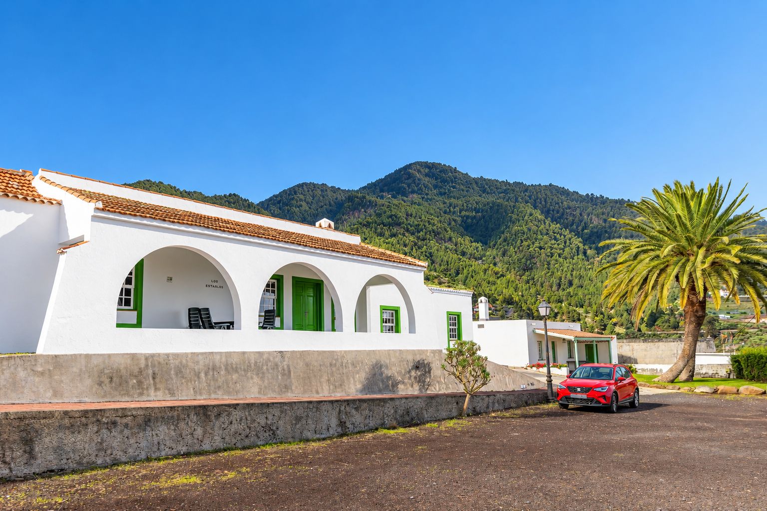 A white building with green doors and windows, a red car parked in front, and a palm tree.
