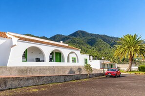 Un edificio blanco con puertas y ventanas verdes, un coche rojo estacionado al frente y una palmera.