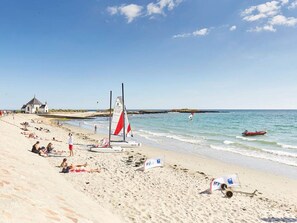 Une plage avec un voilier, des gens qui se détendent et un petit bâtiment en arrière-plan.