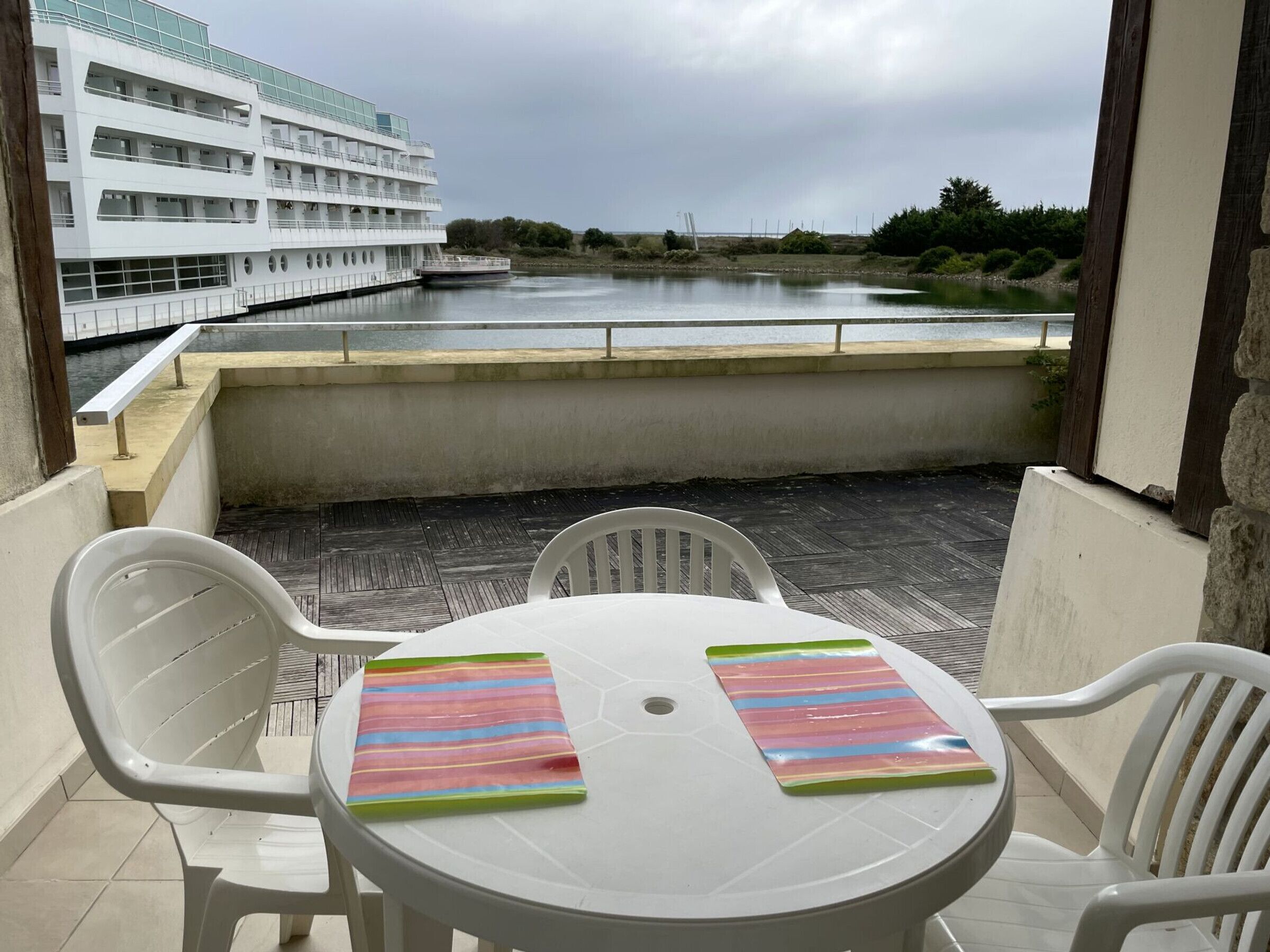 Un balcón con mesa y sillas, con vista a una masa de agua tranquila y un edificio blanco con varios balcones.