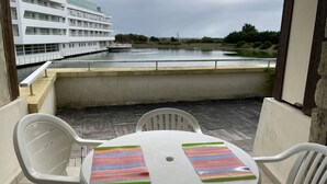 A balcony with a table and chairs, overlooking a calm body of water and a white building with multiple balconies. - 4-person apartment in Arzon, pool and parking, near beach and port (Arzon)
