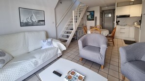 A modern living room with a sofa, armchair, and coffee table. A staircase leads to a dining area with a table and chairs. A kitchen is visible in the background. - T3 Duplex Apartment, 4 Pers, Sarzeau Center, Balcony (Sarzeau)