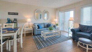 A living room with a coffee bar, a sofa, a dining table with chairs, and a large window. - Oceanfront OIBV 207 in Oak Island, NC: 3 beds, stunning beach views, sleeps 6! (Oak Island)