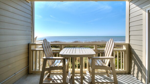A covered outdoor seating area with a table and two chairs, overlooking a beach. - Oceanfront OIBV 207 in Oak Island, NC: 3 beds, stunning beach views, sleeps 6! (Oak Island)