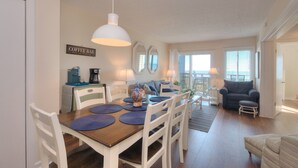 A dining area with a wooden table, white chairs, and a hanging light fixture. There is a coffee bar with a coffee machine and a sign that reads "Coffee Bar." The room has a large mirror, a blue sofa, and a view of the ocean through the windows. - Oceanfront OIBV 207 in Oak Island, NC: 3 beds, stunning beach views, sleeps 6! (Oak Island)
