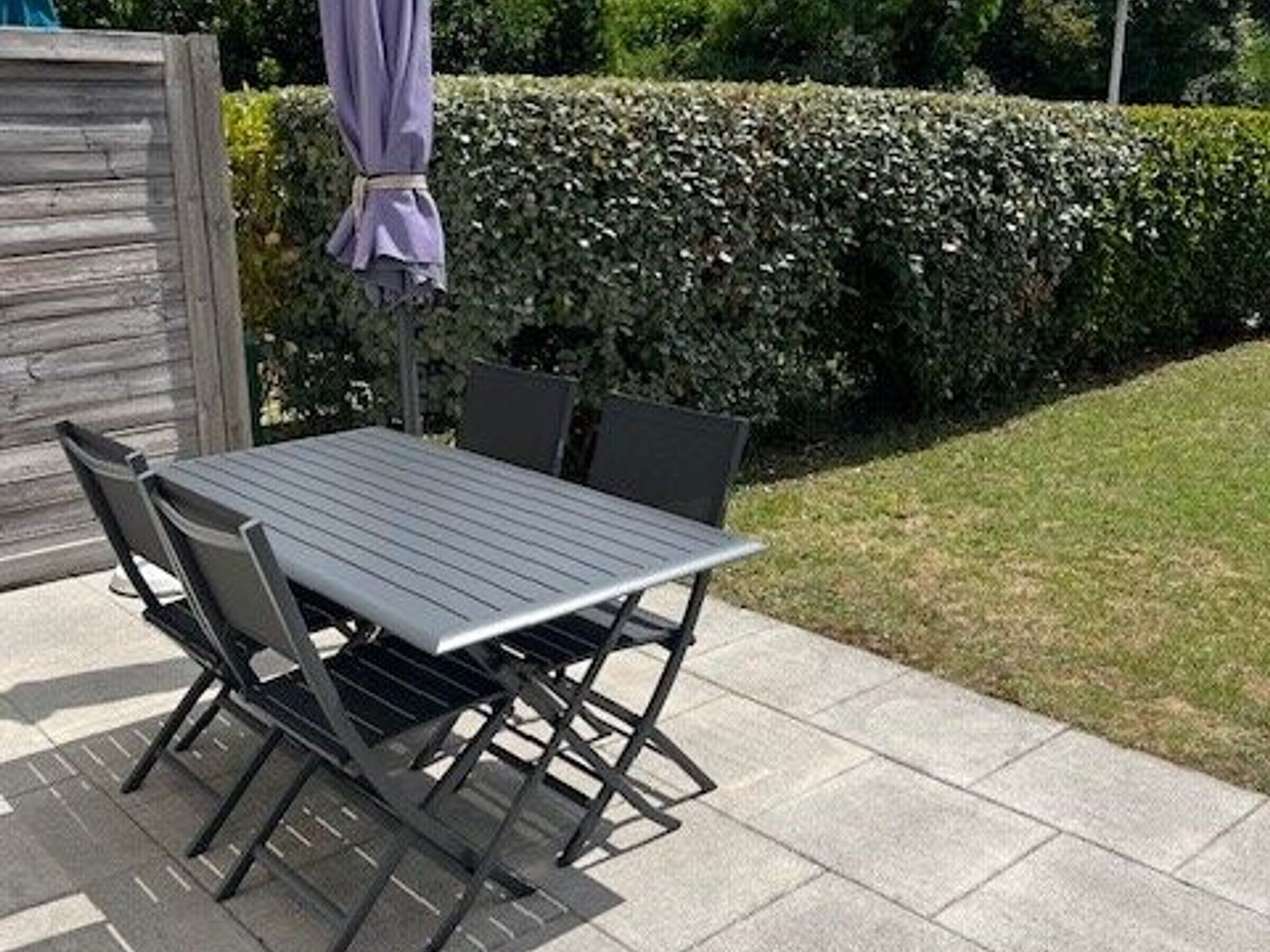 Un patio al aire libre con una mesa y sillas de metal, una valla de madera y un seto al fondo.