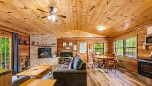 A cabin interior with a fireplace, wooden walls, a dining area, and a television. - New! 3BR*Hot Tub*Game Room*Jacuzzi*Near Dollywood* (Sevierville)