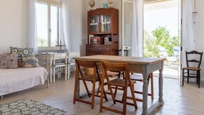 Una habitación amplia con una mesa de comedor de madera, dos sillas, una cama con almohadas estampadas, un armario de madera y vistas a zonas verdes.