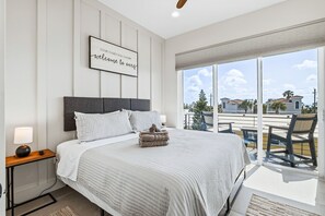 A bedroom with a large bed, a bedside table with a lamp, a framed sign on the wall, and a balcony with chairs and a table.