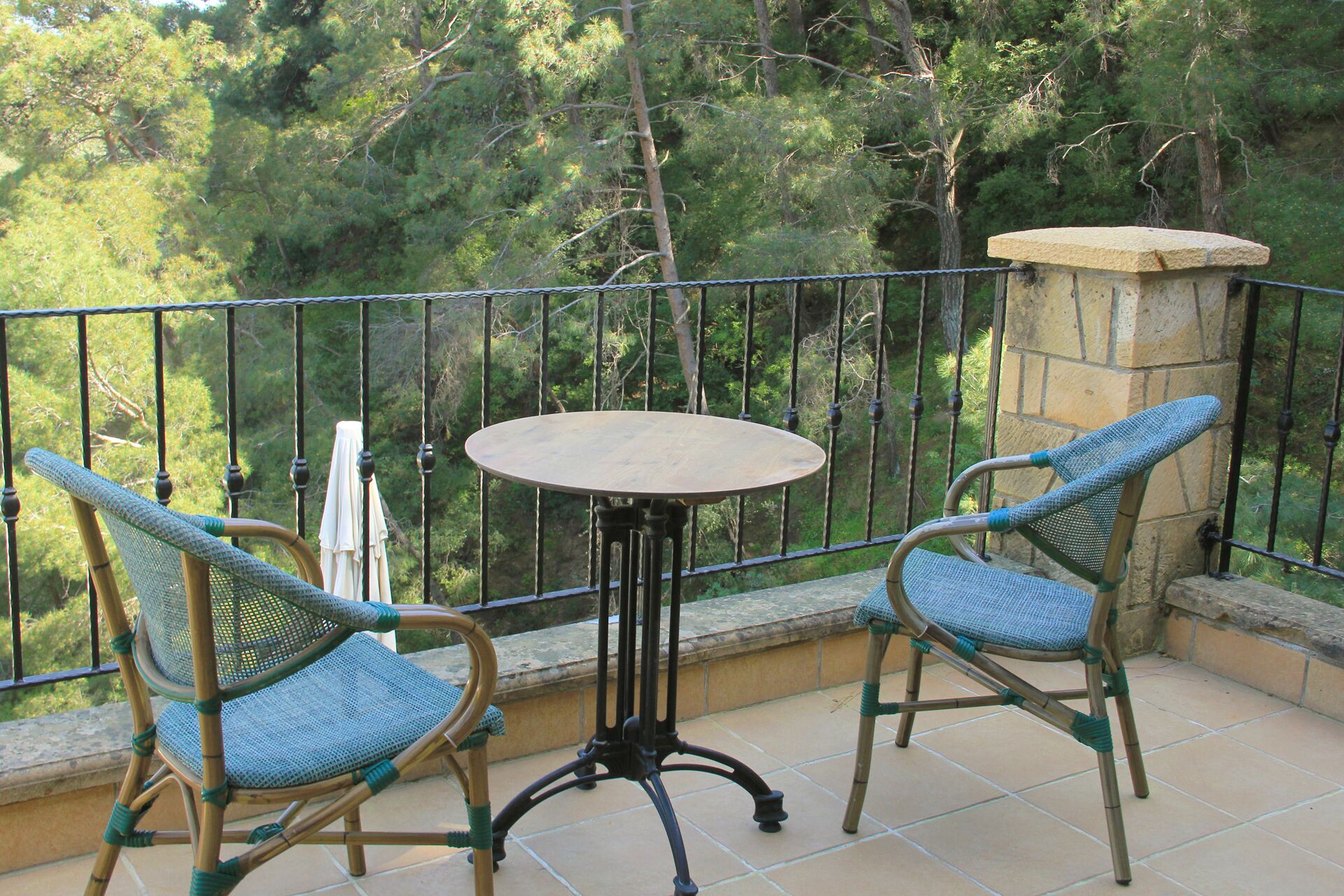 A balcony with two wicker chairs and a small round table, surrounded by a stone pillar and a metal railing, overlooking a forested area.