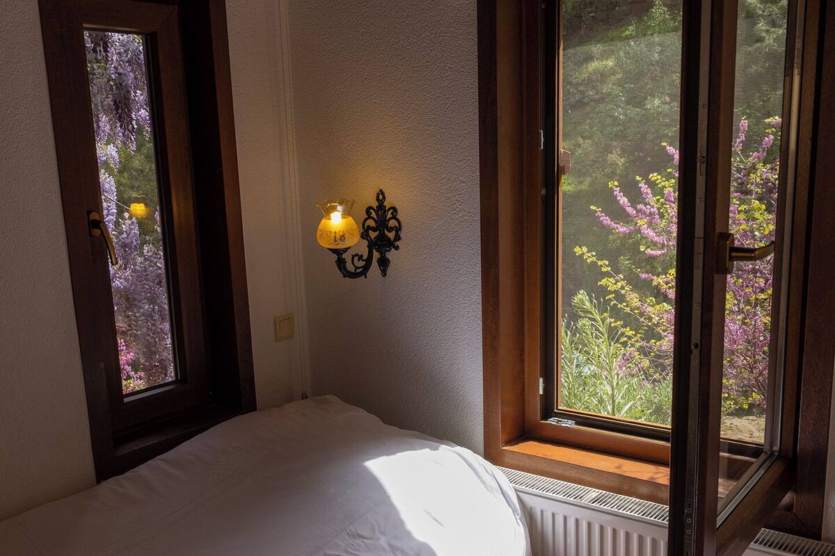 Quarto com duas camas, uma mesa de cabeceira, janela com vista para árvores e luminária fixada na parede.