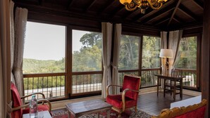 Una stanza con soffitto in legno, ampie finestre con tende, una poltrona rossa, un tavolo e una lampada.