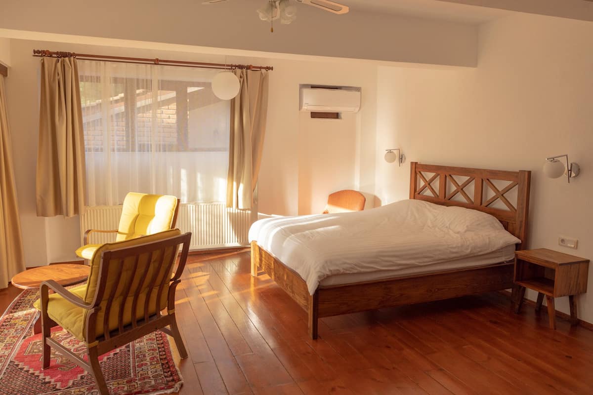Um quarto com cama de madeira, uma cadeira, uma pequena mesa de cabeceira e uma janela com cortinas.