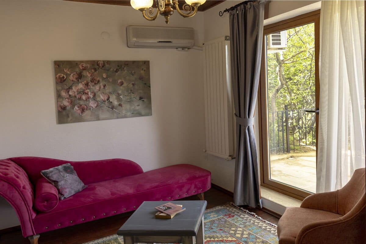 Uma sala de estar com um sofá rosa, uma mesa pequena com um livro, um quadro na parede e vista para um jardim através de uma janela.