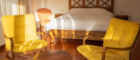 A room with a bed, two yellow chairs, a small round table, and patterned rugs.