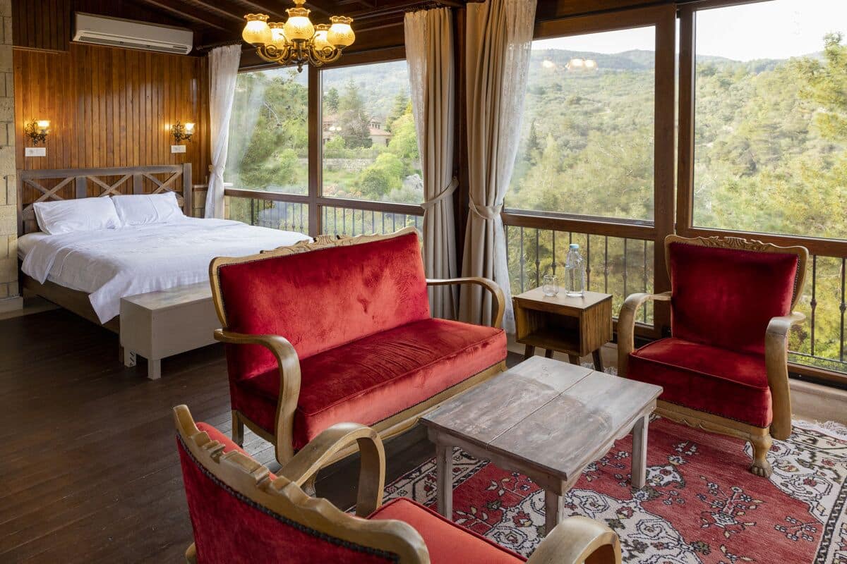 Quarto com uma cama, um sofá vermelho, uma cadeira de madeira, uma mesinha e vista para o exterior.