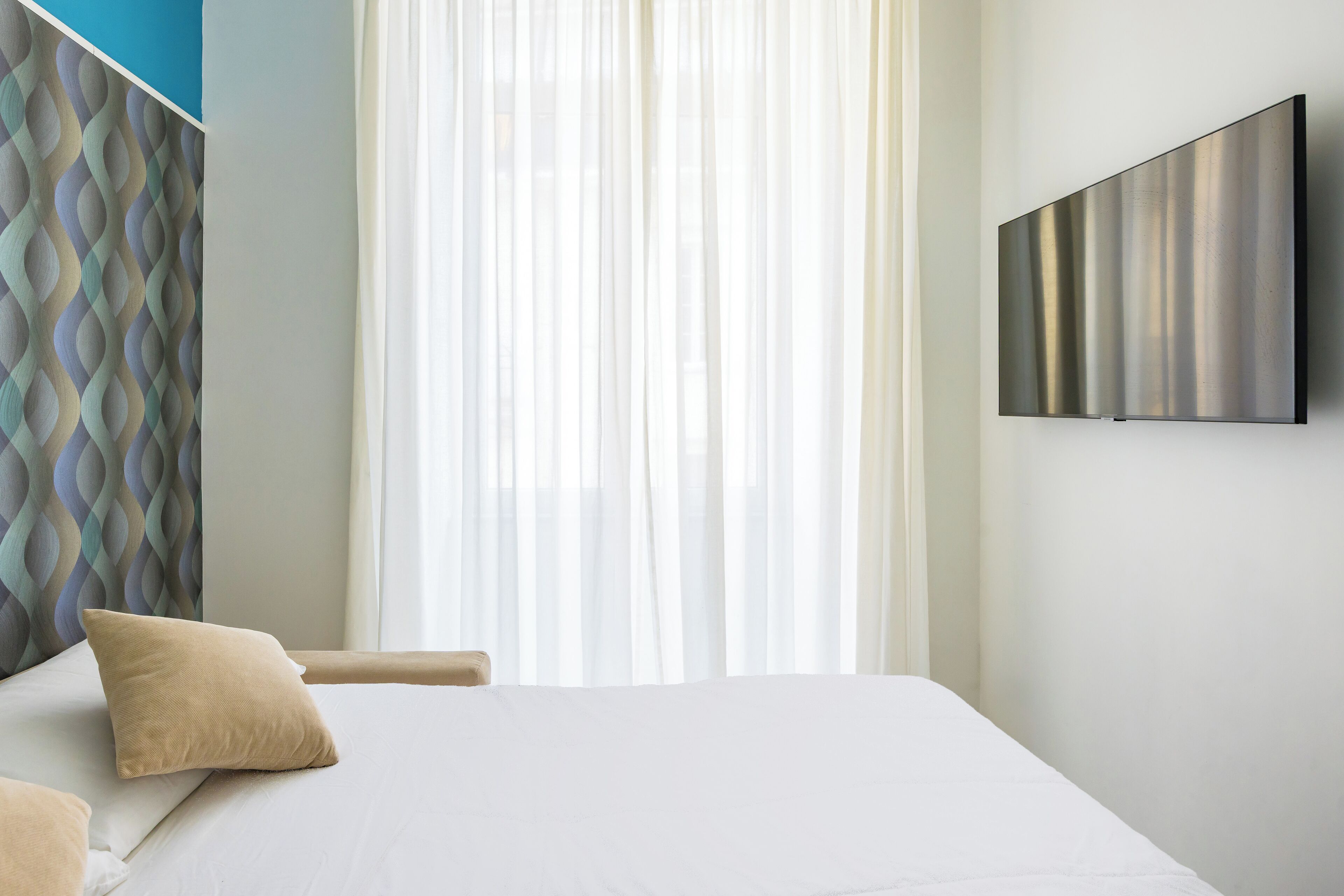 Une chambre d’hôtel avec un lit, un téléviseur fixé au mur et une fenêtre avec des rideaux légers.