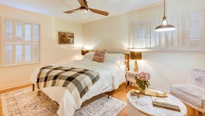 Un dormitorio con cama, ventilador de techo, mesita de noche con lámpara, una silla y una ventana con persianas.