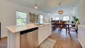 A modern kitchen with a dishwasher, a dining area with wooden chairs, and a large window providing natural light. - Home Near Flagler Beach, Daytona, Parks & More! (Palm Coast)