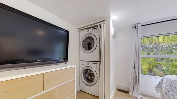 A compact laundry room with a washing machine and dryer, a flat-screen TV mounted above the dryer, and a window with a view of greenery. - Lanai, Pool & Patios: Waterfront Home in Tavernier (Tavernier)