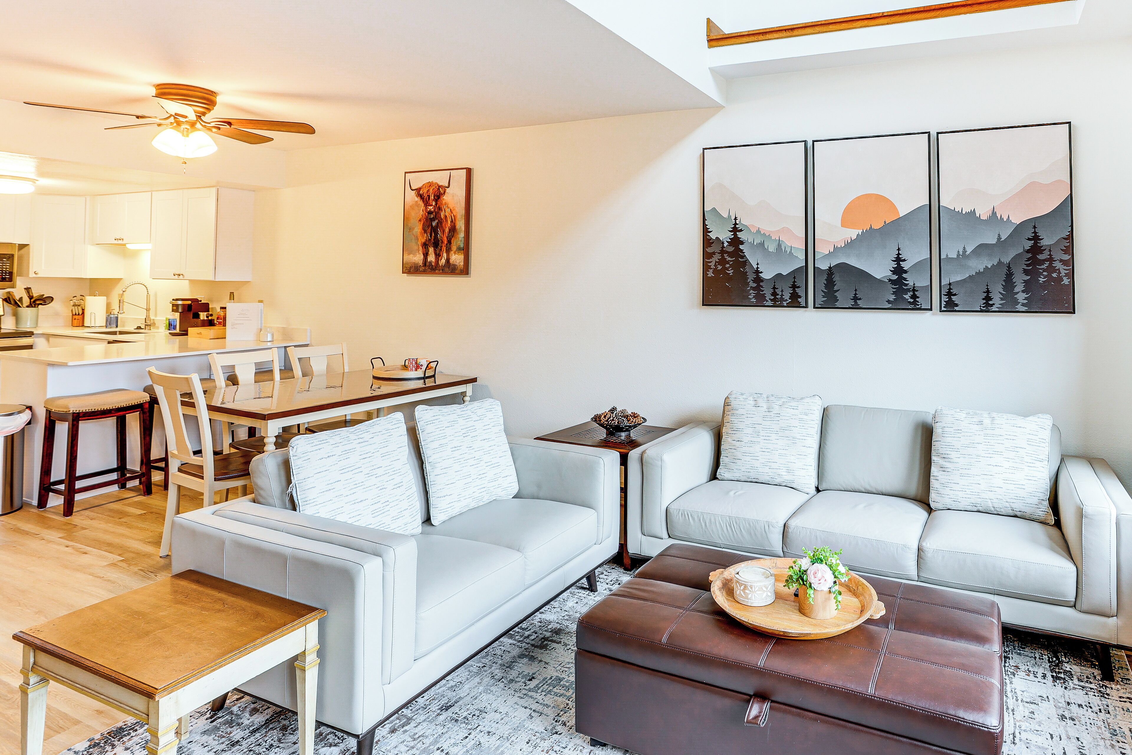 Een moderne woonkamer met een bank, voetenbankje, salontafel en een eethoek met een tafel en stoelen. Er hangen kunstwerken aan de muur en er is een plafondventilator.
