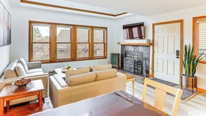 Una sala de estar con chimenea, sofás color beige, una mesa de comedor de madera y ventanas amplias.