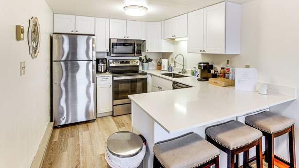 Una cocina moderna con armarios blancos, electrodomésticos de acero inoxidable, una isla central con taburetes y suelo de madera.