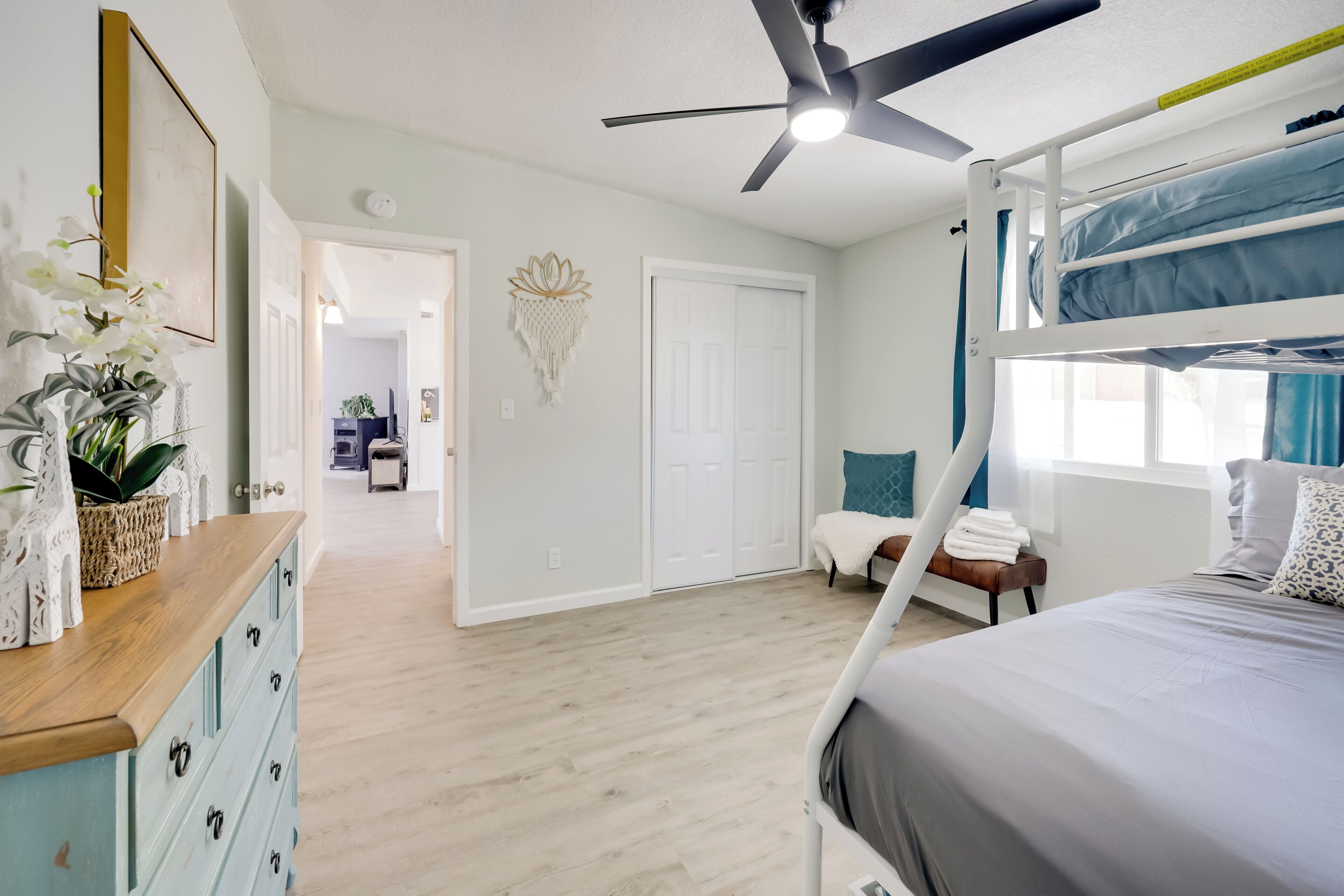Un dormitorio con una cama nido, un ventilador de techo, una ventana con cortinas, una mesita de noche con lámpara y una puerta que da a otra habitación.